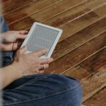Gros plan de mains tenant une liseuse sur un plancher en bois, repr&eacute;sentant un moment de lecture d&eacute;contract&eacute;e.